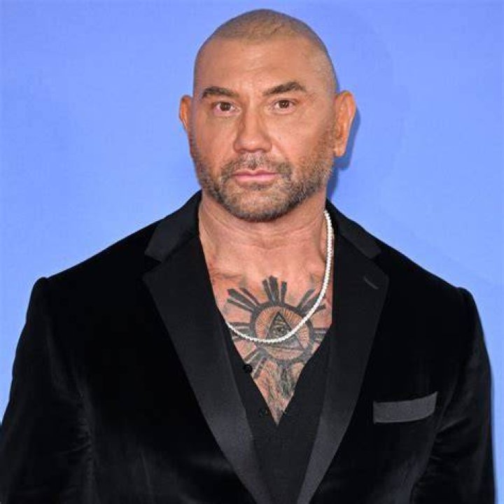 Athlete turned actor Dave Bautista sitting dressed in a leather jacket and dark grey t-shirt with yellow sunglasses in front of a white and grey pin-stripped background.